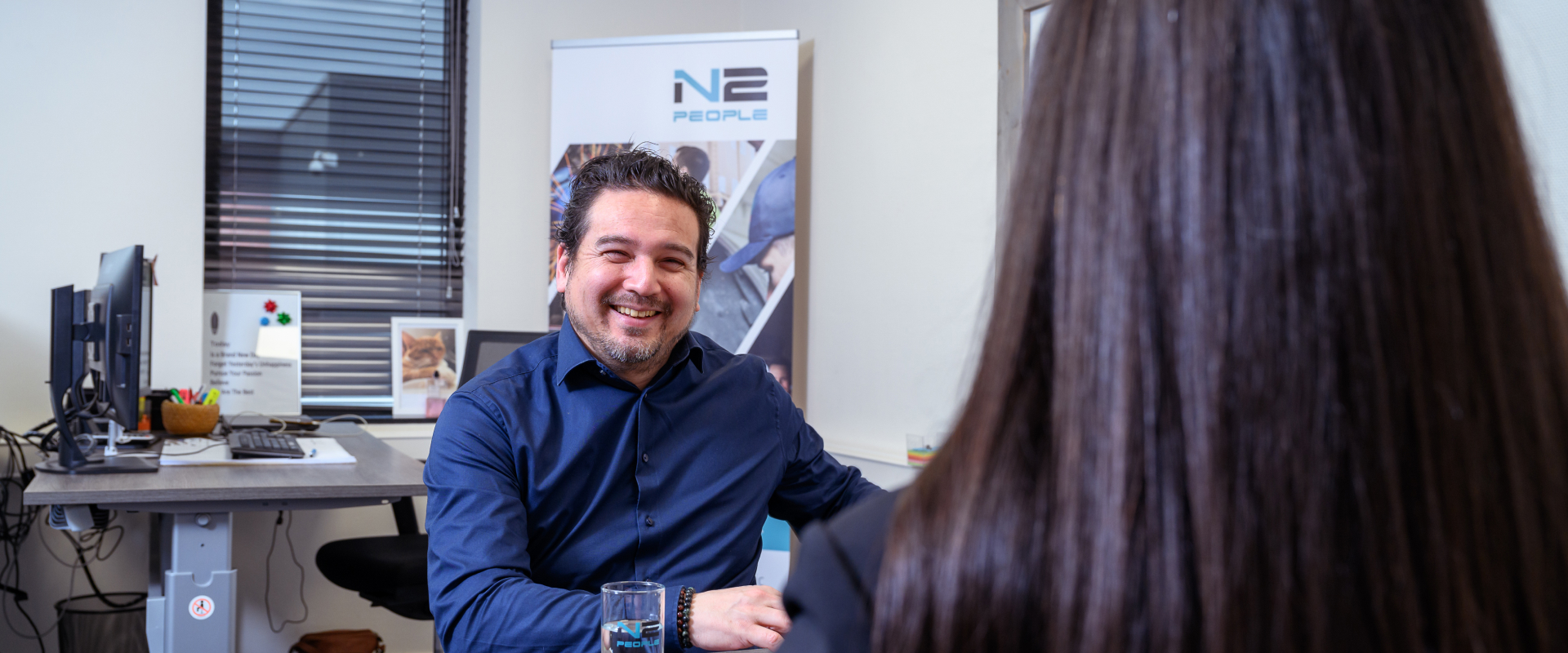 Een lachende man in het N2 People kantoor zit aan een bureau tegenover een vrouw, met een glas water en papieren op de tafel