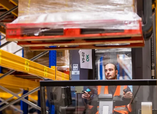 Een magazijnmedewerker in een oranje veiligheidsvest bestuurt een vorkheftruck en plaatst een pallet met goederen op een hoge stelling